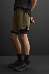 Shorts deportivos color verde oliva con calza interior negra, de tejido técnico ligero y elástico. Cuenta con cintura elástica ajustable con cordón, bolsillos laterales con cierre de cremallera, bajo con aberturas laterales y detalle de logo estampado.