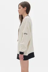 Sweater tejido de punto color crema, con cuello en V con borde negro y doble línea blanca, mangas largas con puños negros, diseño con trenzas en el frente y corte oversize.