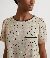Blusa de crepé color beige con estampado de lunares de colores, cuello redondo con ribete negro, manga corta abullonada, bolsillo de parche en el pecho y bajo mullet con aberturas laterales.