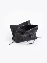 Bolso tote negro de cuero vacuno con textura granulada. Presenta cierres funcionales en los laterales para expandir el espacio y dos asas de mano. Incluye un tag colgante con aplique metálico y un tirador de cremallera con forma de figura.