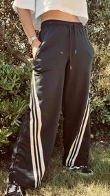 Pantalón de jogging color gris claro, corte ancho, con cordón ajustable en la cintura y tres rayas negras verticales en los laterales.
