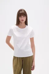 Remera blanca de algodón con cuello redondo y manga corta. Presenta un bordado lineal en tono claro, simulando formas orgánicas o florales, adornado con pequeñas mostacillas.