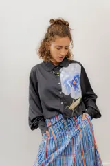 Camisa corta de seda negra con cuello clásico, cierre frontal con botones y mangas largas abullonadas. Presenta un estampado de flor y un insecto en el frente.