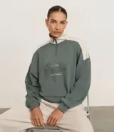 Buzo deportivo de modal, color verde grisáceo, con cuello alto color beige y cierre parcial de cremallera. Presenta un diseño de bloques de color con hombros en beige y un estampado central circular con la palabra "Monaco".