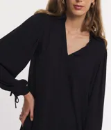 Vestido corto negro de viscosa con cuello en V, mangas largas abullonadas con lazo en los puños y falda con volantes.