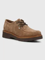 Zapato bajo de mujer color camel, estilo mocasín o derby, confeccionado en gamuza. Presenta suela gruesa de plataforma color marrón oscuro con costura visible en color claro. El sistema de ajuste es mediante cordones marrones que pasan por ojales metálicos tipo gancho.