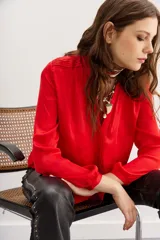 Camisa roja de manga larga con cuello redondo y tajo frontal en el escote. Presenta botones ocultos al frente, puños con botón, alforzas en los hombros y frunces.