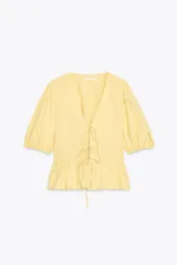 Top amarillo de escote pico y manga corta abullonada con elástico. Detalle de combinación de tejidos a tono y bajo con volante. Cierre frontal con lazadas.