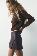 Falda pantalón corta marrón de tiro medio con efecto piel, falsos bolsillos de vivo en la espalda y cierre lateral con cremallera oculta.