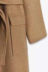 Abrigo largo color camel confeccionado en hilatura con lana. Tiene cuello clásico, cierre frontal con botones ocultos, bolsillos de parche y cinturón del mismo tejido.