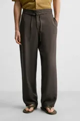 Pantalón marrón oscuro de corte amplio, con cintura ajustable con cordón, bolsillos plastrón delanteros y traseros, detalle de costuras frontales y cierre frontal con cremallera y gancho metálico oculto.