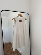 Vestido blanco sin mangas, con escote redondo y detalle de pliegues verticales en el frente, con lazos ajustables en el escote. Corte amplio y holgado.