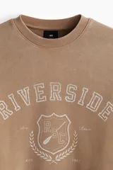 Buzo de tejido pesado de algodón con efecto lavado, color marrón topo. Presenta un diseño estampado en el frente con el texto "RIVERSIDE SOCIAL CLUB" y un escudo con un remo. Tiene cuello redondo, hombros caídos y puños y bajo acanalados.