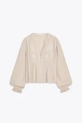 Blusa color beige de manga larga con volantes en los puños, cuello mao y bordados florales en el frente.