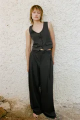 Pantalón de vestir gris oscuro, de tiro alto y corte palazzo. Presenta trabillas, bolsillos laterales y traseros de vivo, pliegues frontales y cierre frontal con cremallera, botón interior y ganchos metálicos.