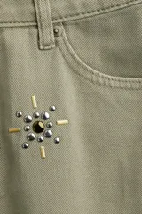 Pollera corta de denim color verde oliva, estilo cinco bolsillos, con cierre y botón frontal. Presenta apliques de tachas metálicas doradas y plateadas en forma de estrella o brújula distribuidas en el frente.