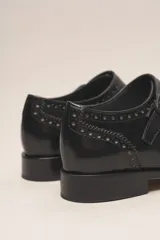 Zapato bajo de cuero negro, estilo monk strap con dos hebillas ajustables sobre el empeine. Presenta detalles de brogue (perforaciones) en la puntera y alrededor del borde superior. Tiene un taco bajo y ancho.
