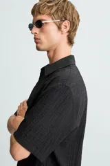 Camisa de hombre de manga corta, cuello solapa y corte relaxed fit. Tejido blanco con textura sutilmente arrugada o plisada. Cierre frontal de botonadura y bajo con aberturas laterales.