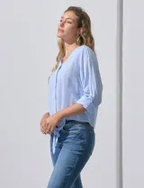 Blusa de algodón a rayas verticales finas en tonos blanco y lila, con escote en V, abertura frontal con botones y mangas largas regulables con presilla y botón. El ruedo termina anudado al frente.