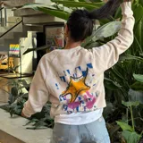 Buzo color beige de algodón y poliéster, con cuello redondo y puños acanalados. Presenta un pequeño estampado en el frente con una estrella de mar amarilla sobre un fondo a cuadros violeta y celeste.