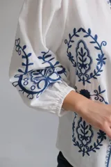 Camisa blanca de manga larga con elástico en puños, cuello mao con botones en la parte delantera y bordados florales azules en el frente y las mangas.