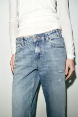 Jeans de tiro bajo, corte recto y pierna ancha, color celeste claro desgastado. Presenta roturas visibles en la zona de la rodilla y pequeños desgastes en los muslos y bolsillos.
