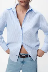 Camisa celeste y blanca a rayas verticales, de corte recto, con cuello solapa, escote pico y manga larga. Cierre frontal con botones.