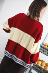 Cardigan de punto grueso con diseño a rayas horizontales en color crema, rojo y marrón. Presenta cuello redondo, cierre frontal con botones oscuros y ribetes a rayas marrones y celestes en cuello, puños y ruedo. Lleva un pequeño bordado con el logo de la marca en el pecho.