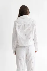 Campera blanca de lino con bordado calado, corte clásico de campera de jean, con cuello camisero, cierre frontal con botones de pasta color marfil, bolsillos de parche con solapa en el pecho y mangas largas con puños abotonados.