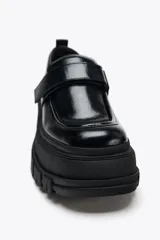 Zapato tipo mocasín negro con plataforma y suela track. Presenta tira ajustable adherente y tirador trasero.