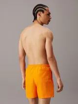 Bañador corto color naranja con cintura elástica ajustable con cordón blanco, bolsillos laterales y logo de Calvin Klein estampado en blanco en la pierna izquierda.