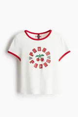 Remera blanca de algodón con cuello y mangas con ribete rojo. Estampado con cerezas y texto "Ibiza" en rojo.