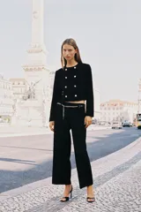 Blazer corta negra de cuello redondo y manga larga, con cierre frontal cruzado con botones dorados y falsos bolsillos delanteros.
