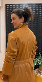 Gabardina color mostaza con cuello camisero, cierre frontal con botones y cinturón con hebilla.