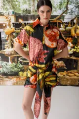 Vestido camisero corto con estampado abstracto en tonos naranja, rosa, amarillo y negro. Tiene cuello camisero, mangas cortas y cierre frontal con botones. Incluye lazo en la cintura para ajustar.