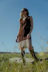 Vestido corto marrón de gamuza sintética, sin mangas, con escote en V y detalle de flecos en el ruedo. Presenta apliques bordados en el frente y laterales.