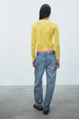 Chaqueta de punto amarilla con cuello redondo, manga larga y cierre frontal con botones negros. Presenta ribetes rosas en cuello, puños y bajo.