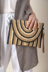 Sobre de mano de rafia con diseño de rayas horizontales en tonos beige, dorado y negro. Tiene cierre superior y correa de cuero sintético marrón.