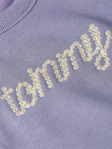 Sweater de felpa color lila claro, con cuello redondo y puños y dobladillo acanalados. Presenta el logo 'Tommy' bordado en el pecho con apliques de flores blancas y amarillas.
