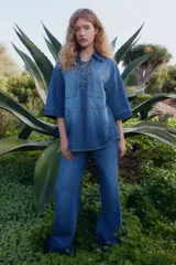 Camisa popover de denim azul con costuras en color contrastante, corte oversized, hombros caídos, cuello abierto con detalle de cordón, bolsillos grandes superiores, mangas al codo y bajo levemente redondeado.