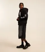 Saco tipo hoodie con cierre frontal de cremallera, color negro, corte boxy y capucha. Presenta estampa de letras en blanco en ambas mangas y un colgante de cruz metálico en el tirador de la cremallera principal.