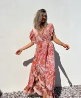 Vestido largo de corte evasé con estampado floral en tonos rosados y anaranjados. Tiene escote en V cruzado, mangas cortas con volantes y falda con volantes asimétricos.