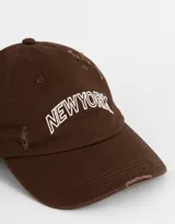 Gorra de béisbol marrón con bordado frontal de la leyenda "New York" en blanco. Presenta detalles de roturas en la visera y la corona.