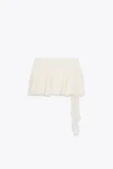 Falda pantalón corta color blanco roto, de tiro bajo, con cintura drapeada y bajo acabado en volantes superpuestos. Presenta un lazo largo de tela que cae desde el lateral.