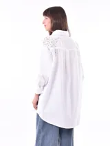 Camisa blanca de corte oversize con cuello clásico, cierre frontal con botones y mangas largas con puños abotonados. Presenta detalle de encaje en los hombros.