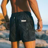 Short de baño negro con estampado de hojas de palmera en blanco. Tiene elástico en la cintura, cordón ajustable, tres bolsillos y mesh interior hipoalergénico.