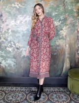 Vestido camisero midi de gasa estampada con flores rojas y blancas sobre fondo marrón. Tiene cuello con solapa, abertura central con botones, mangas largas y lazo de ajuste en la cintura.