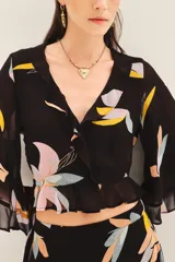 Camisa crop de manga larga con volados en mangas y cintura, confeccionada en viscosa georgette estampada con motivos florales abstractos en tonos pastel sobre fondo negro.