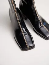 Botas de cuero charol negro con caña media y punta cuadrada. Cuentan con cierre trasero, tirador y bordado contrastante en el empeine. El taco es de madera y la suela de goma.