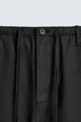 Pantalón negro de corte relajado y pierna ancha, confeccionado en mezcla de algodón y lino. Presenta cintura elástica ajustable con cordón y cierre frontal con cremallera y botón.
